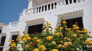 A white building with balconies and shutters, surrounded by yellow flowers. - Binibeca Mar (Sant Lluis)