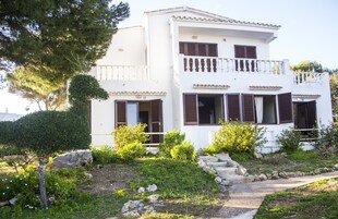 A two-story white building with brown shutters, a balcony, and a stone pathway leading to the entrance.