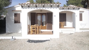 A white building with two doors, each with a wooden shutter. There are two wooden tables and chairs in front of each door. The building has a tiled roof and is surrounded by gravel. - Binibeca Mar (Sant Lluis)