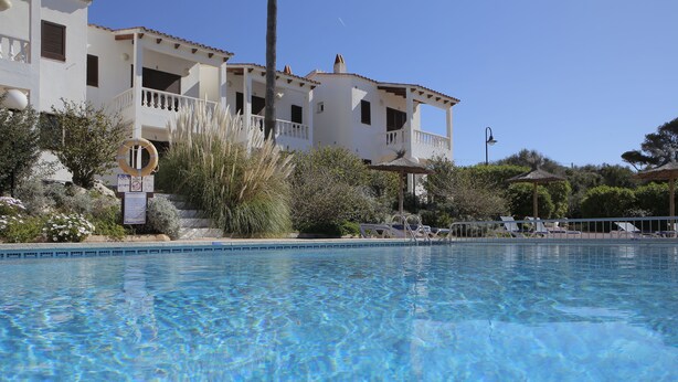 A clear swimming pool with a view of a white building with balconies and a lifebuoy sign. - Binibeca Mar (Sant Lluis)