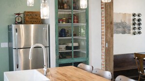 Una cocina con un refrigerador de acero inoxidable, una mesa de madera con sillas, un armario de vidrio con marco verde y botellas, y lámparas colgantes.