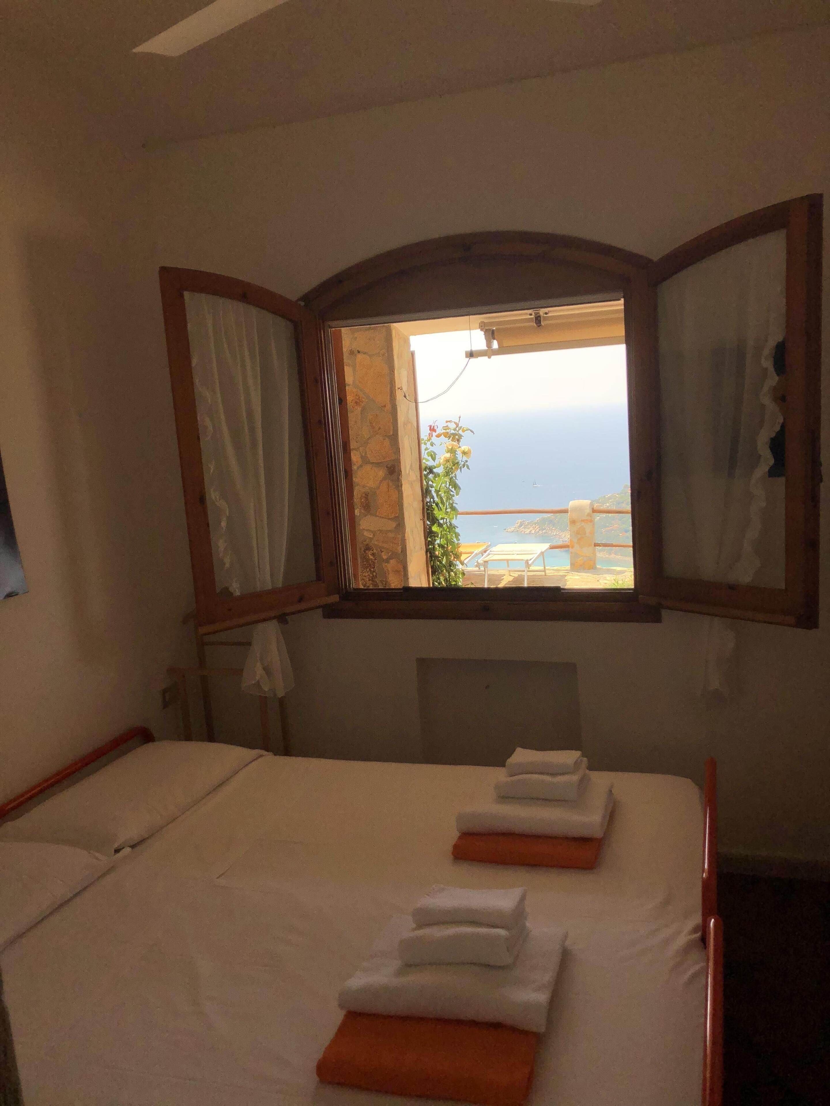 A bedroom with a bed, a window with curtains, and a view of the sea.
