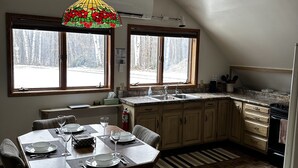 Ein Essbereich mit einem Tisch für vier Personen, eine Küche mit Herd und Spüle sowie ein Blick auf eine verschneite Landschaft durch große Fenster.
