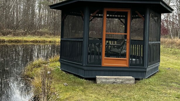 Ein Pavillon mit Glastür und kleiner Veranda, umgeben von einer Rasenfläche und einem Gewässer, mit Bäumen im Hintergrund.