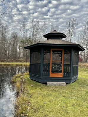 A gazebo with a glass door and a small porch, surrounded by a grassy area and a body of water, with trees in the background.