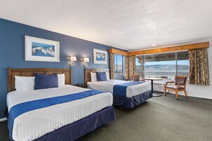A hotel room with two beds, a small table, and a chair. There are two windows with curtains, and a painting on the wall.