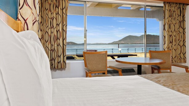 A hotel room with a large bed, two chairs, a table, and a view of the sea and mountains through large windows. - Skylark Shores Resort (Lakeport)