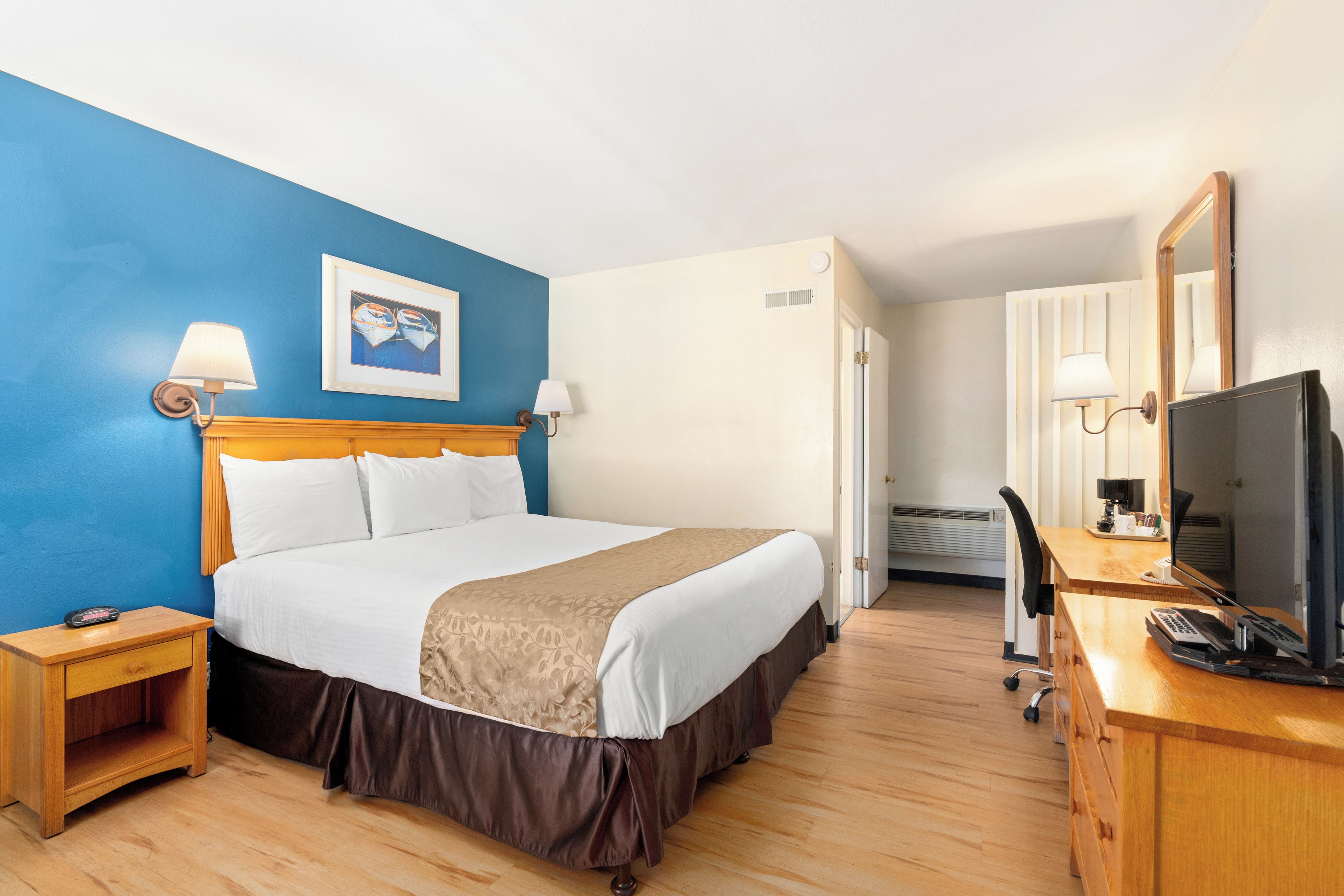 Quarto de hotel com cama, mesas de cabeceira, escrivaninha com computador, televisão e espelho.