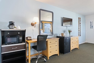 A hotel room with a wooden desk, a black office chair, a microwave, a coffee maker, a dresser, a mirror, and a television.