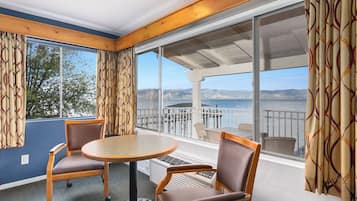 A room with a round table and two chairs, large windows with curtains, and a view of the sea and mountains.