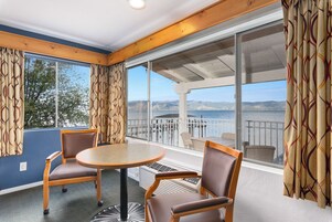 A room with a round table and two chairs, large windows with curtains, and a view of the sea and mountains.