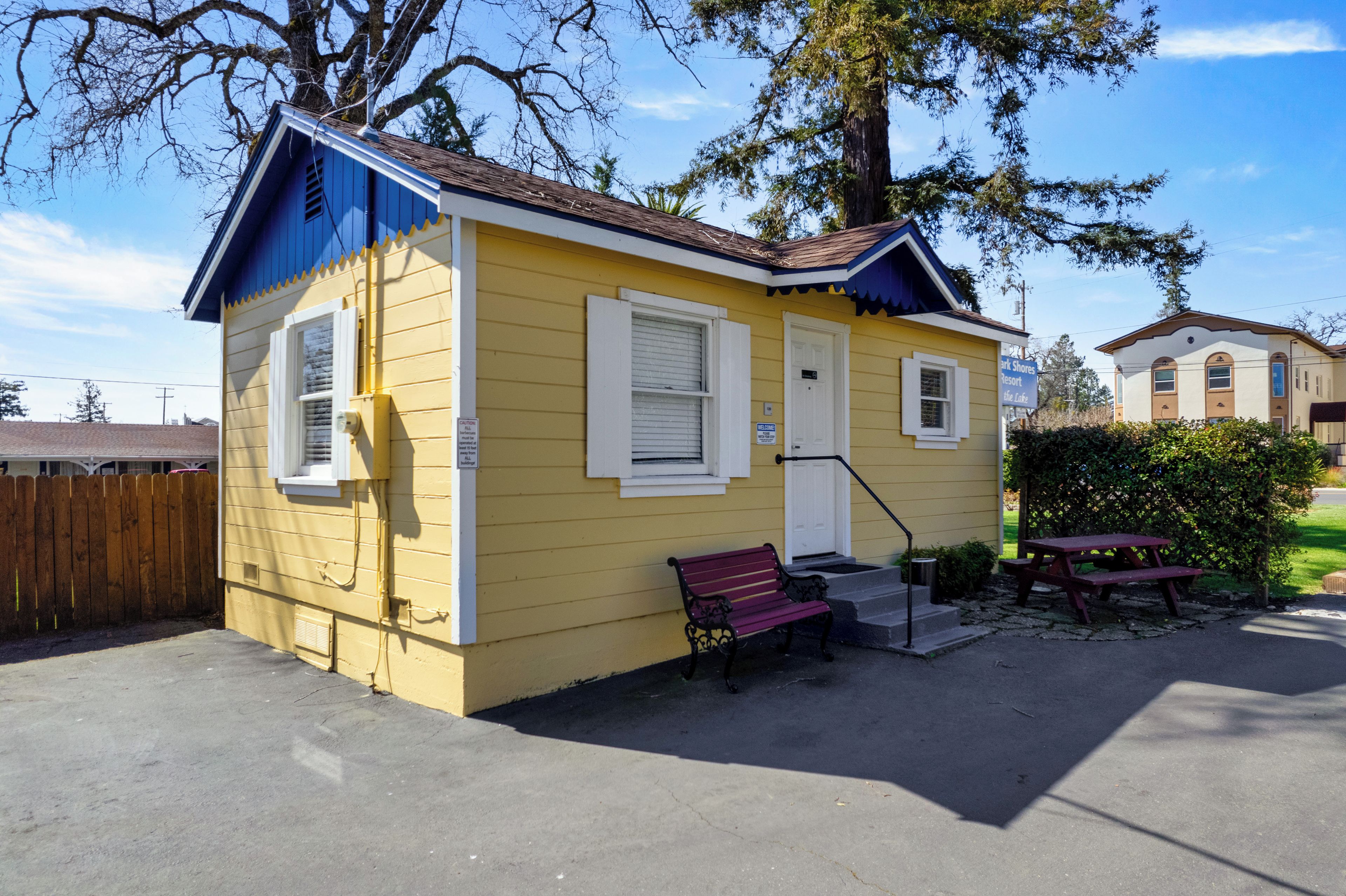 Un edificio amarillo con techo azul, cerca de madera, un banco y una mesa de picnic.
