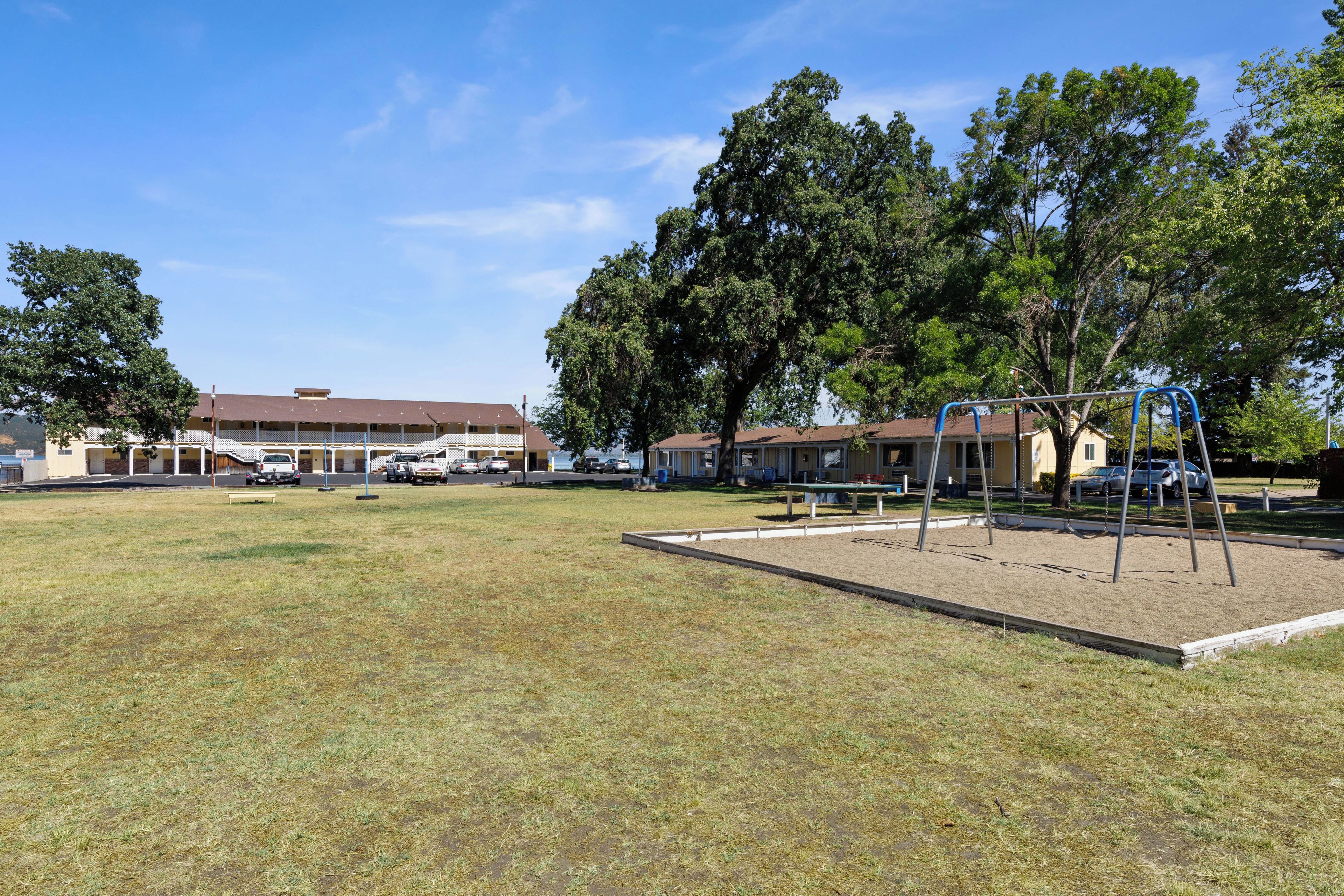 A playground with swings and a sandbox, surrounded by trees and a building with a brown roof.