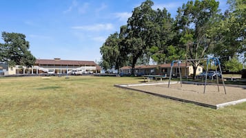 Um parquinho com balanços e um caixa de areia, cercado por árvores e um edifício com telhado marrom.