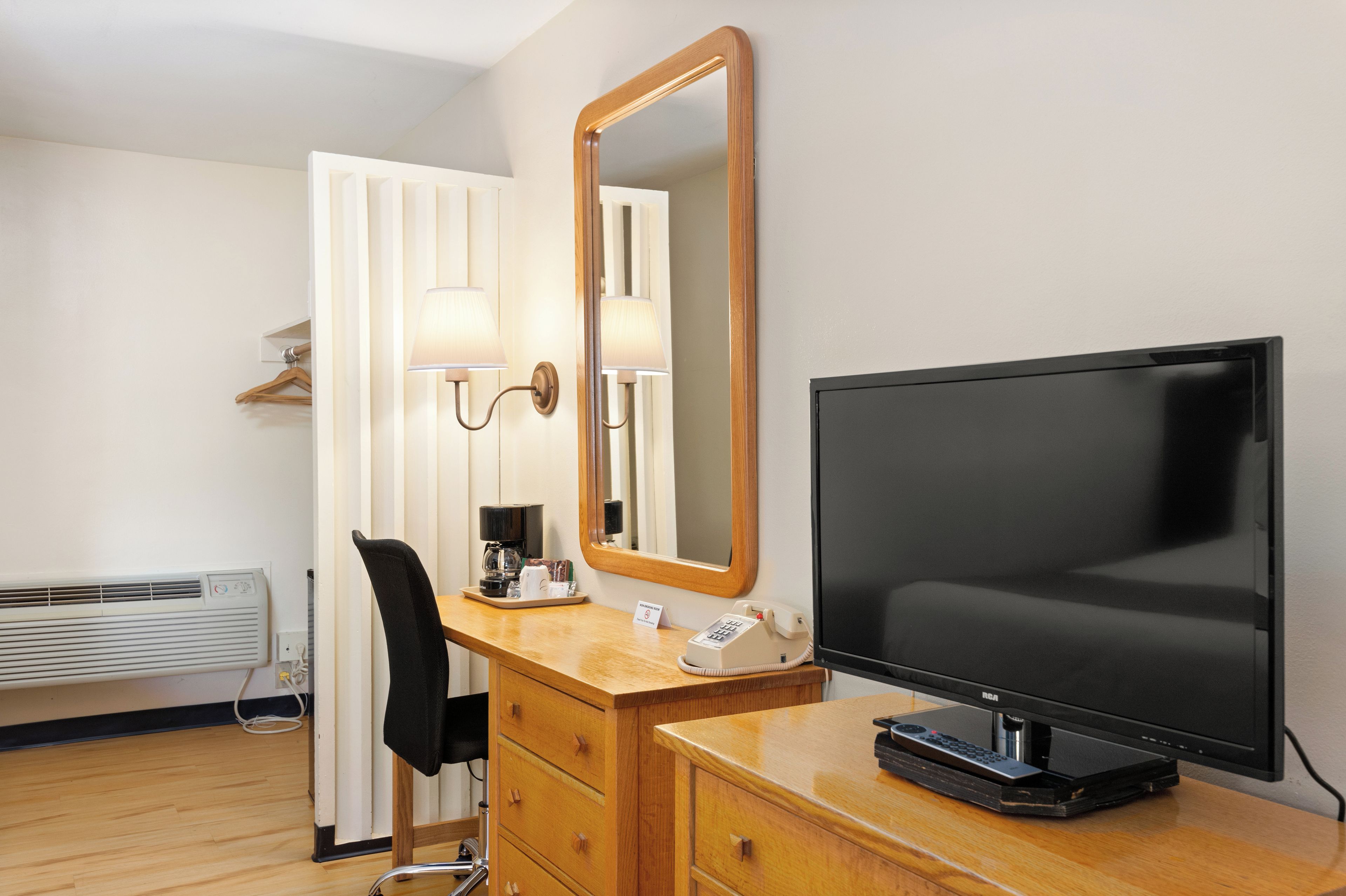 A hotel room with a wooden desk, a mirror, a television, a chair, a telephone, and a heater.