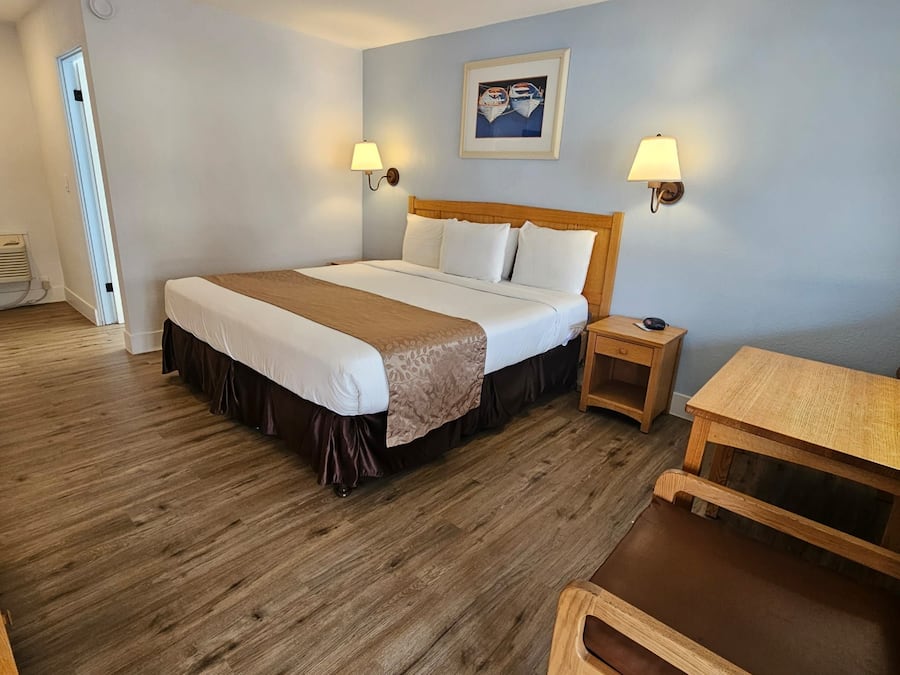 Ein Hotelzimmer mit einem großen Bett, zwei Nachttischlampen, einem Holznachttisch, einem kleinen Holztisch und einer Tür zu einem weiteren Raum.