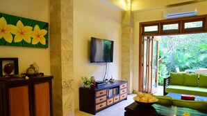 Een woonkamer met een aan de muur bevestigde televisie, een houten kast, een groene bank en een eettafel met een fruitschaal.