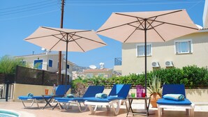 Ein Poolbereich mit Liegestühlen und Sonnenschirmen, einem gefliesten Boden und Gebäuden im Hintergrund.