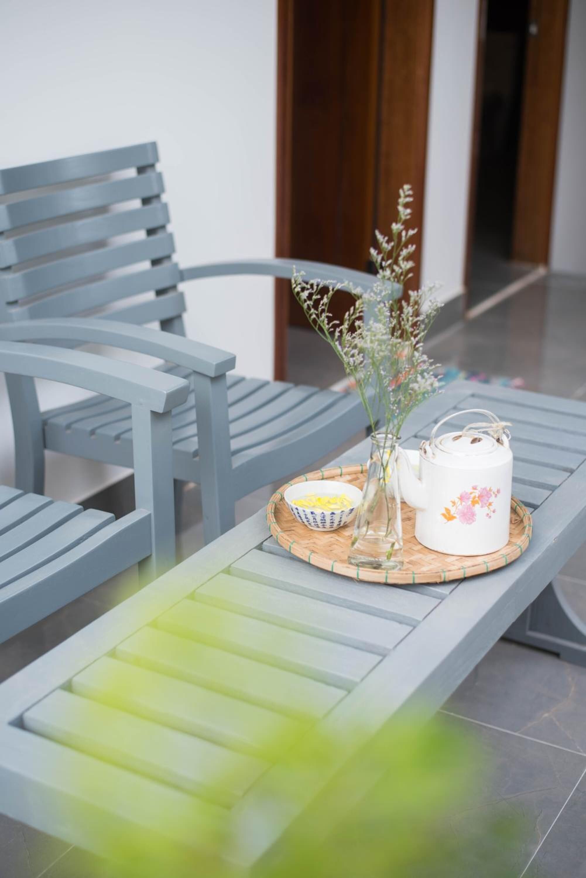 A modern outdoor seating area with a grey metal bench and a table. On the table, there is a white teapot with floral designs, a glass vase with flowers, and a small bowl with yellow items.