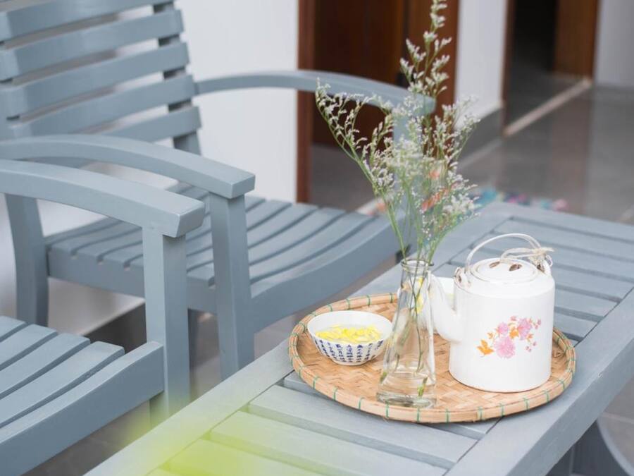 Ein moderner Außenbereich mit einer grauen Metallbank und einem Tisch. Auf dem Tisch stehen ein weißer Teekessel mit floralen Mustern, eine Glasvase mit Blumen und eine kleine Schale mit gelben Gegenständen.