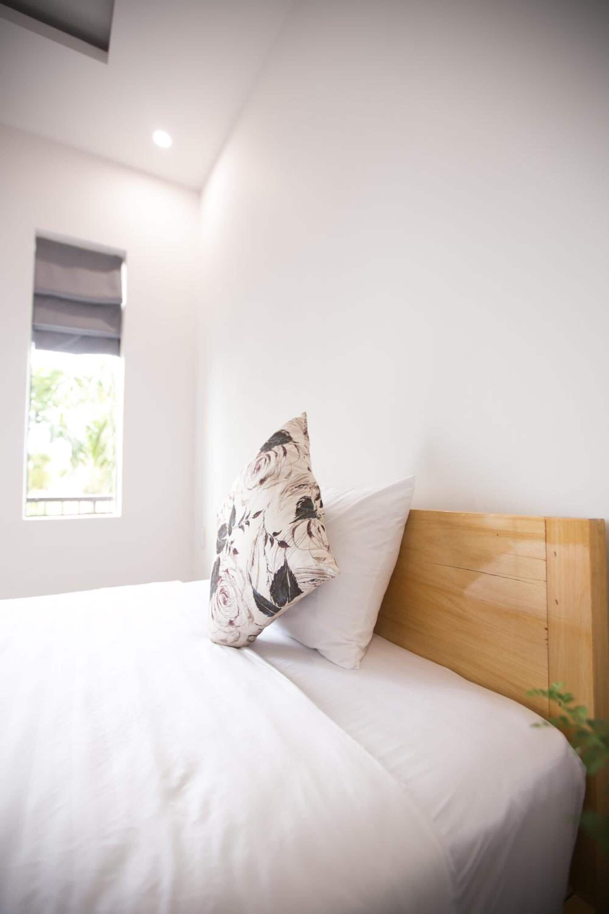 A neatly made bed with white bedding and a wooden headboard, a window with a view of greenery, and a single potted plant on the right.