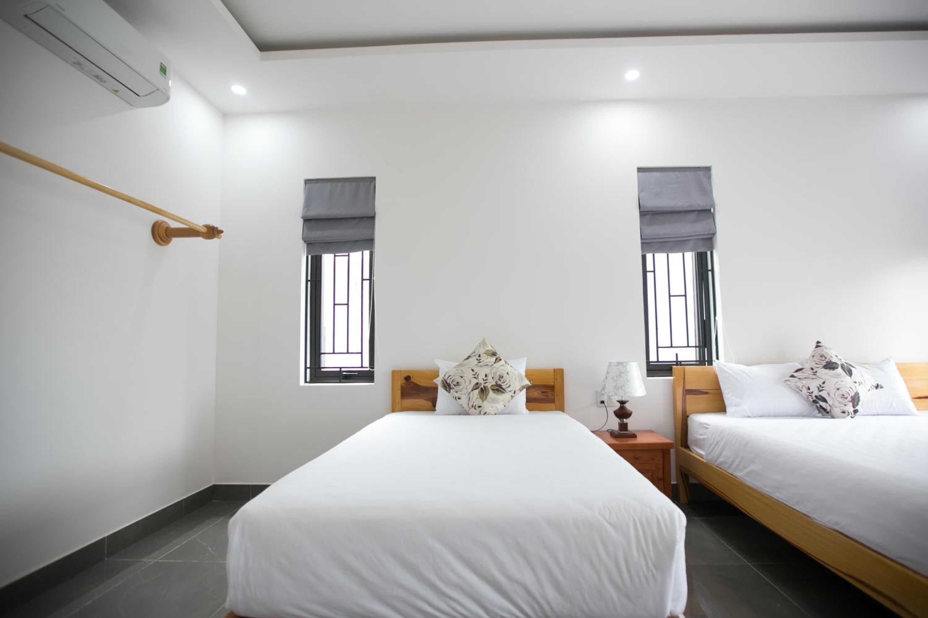 Habitación de hotel con dos camas, cabecero de madera, mesita de noche con lámpara y ventana con persiana.