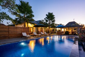Una piscina con sillones de descanso y sombrillas, rodeada de cercas de madera y palmeras.