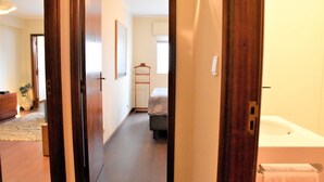 A hotel room with a wooden door, a bed with a white mattress, a bedside table, a hanging chair, and a bathroom with a bathtub and toilet. - Ribeira Oporto Apartment (New) (Porto)
