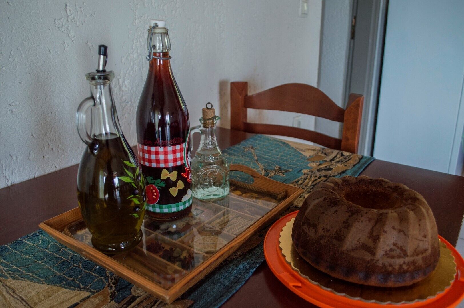 A dining table with a bottle of olive oil, a bottle of balsamic vinegar, and a bottle of water. There is a wooden tray with a cake on an orange plate.