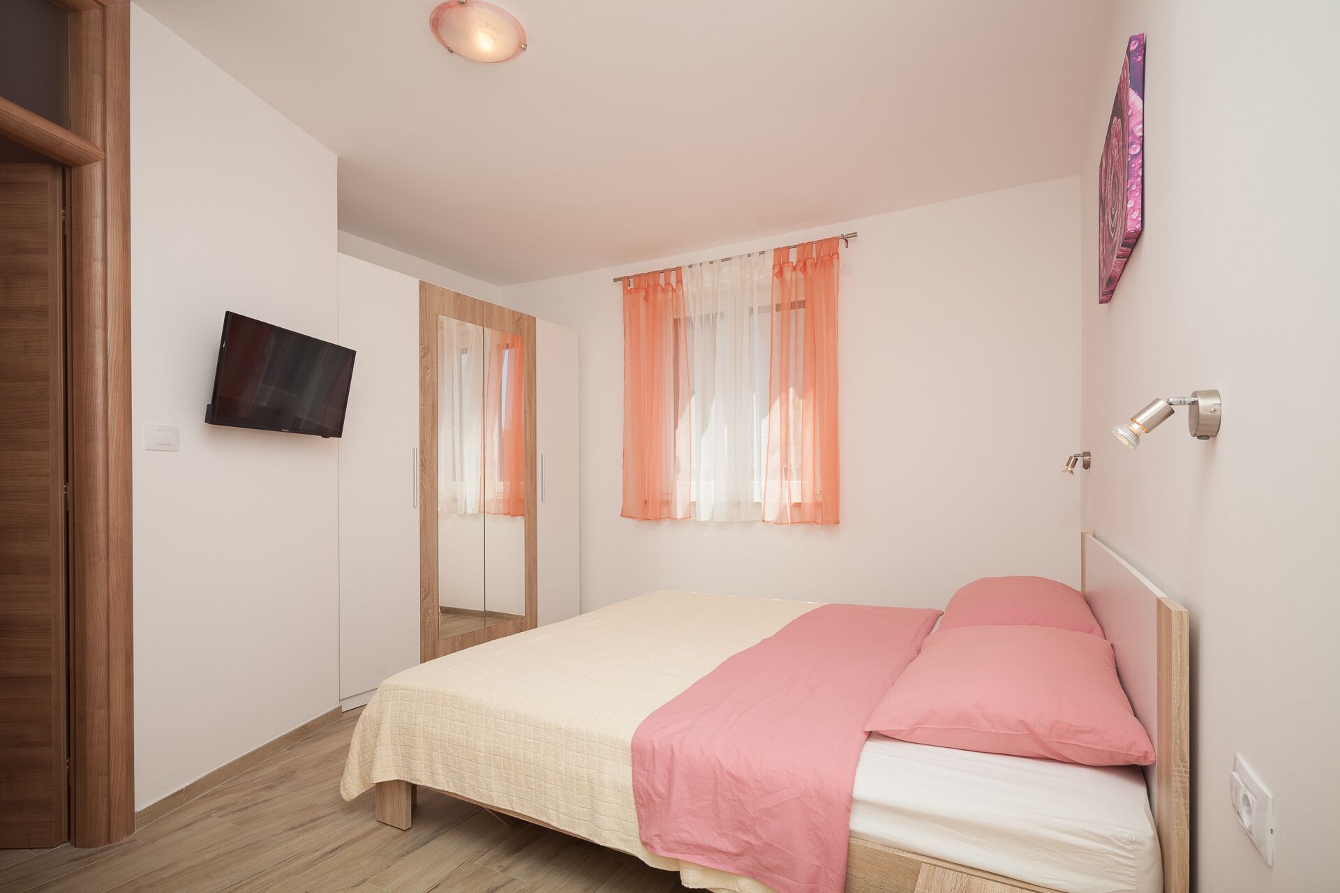 Un dormitorio con una cama, un televisor montado en la pared y un ventanal con cortinas translúcidas.