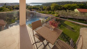 A balcony with a glass railing, wicker furniture, and a view of a swimming pool and greenery.