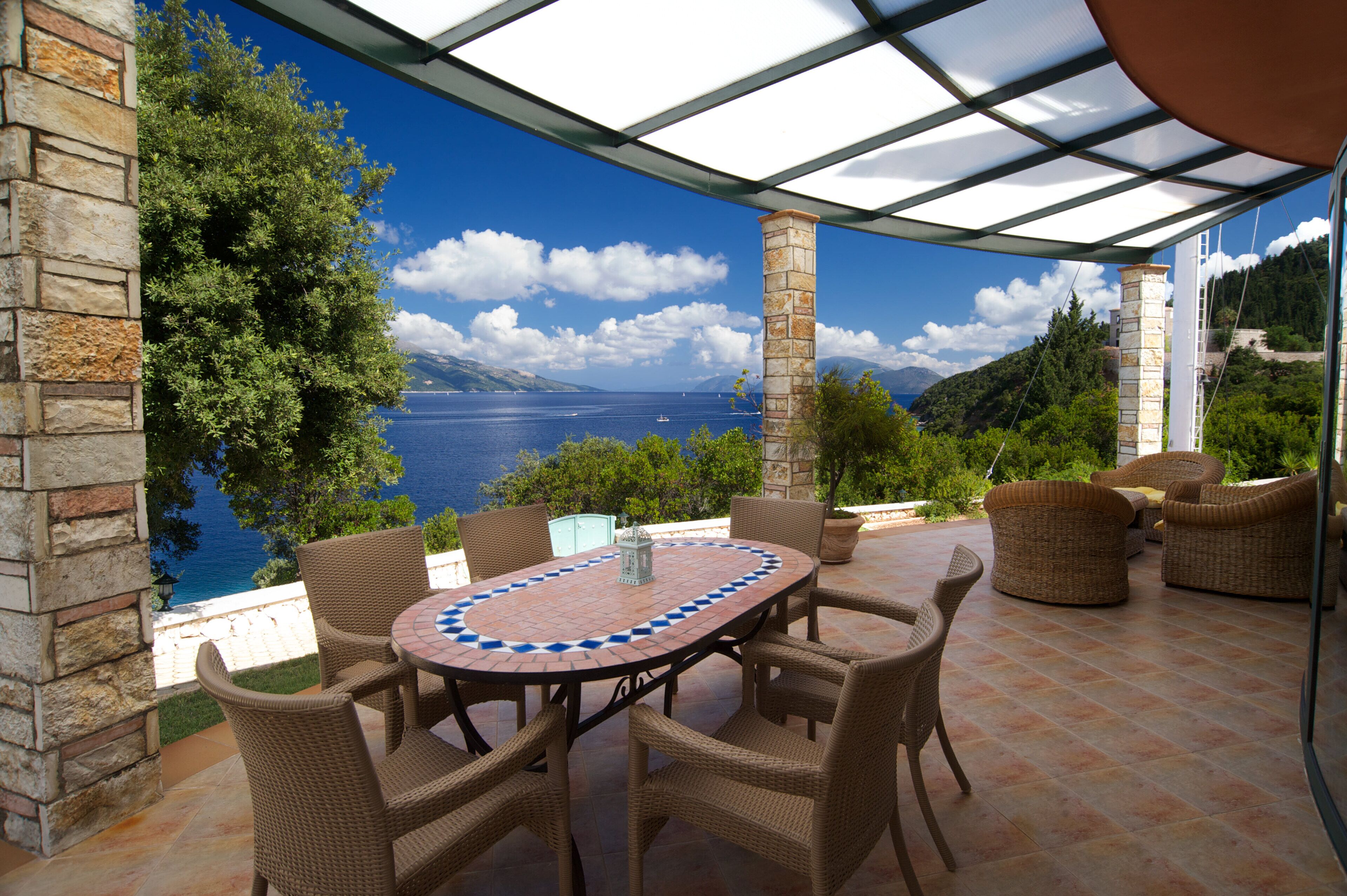 Une terrasse extérieure aménagée avec des meubles en osier et une table carrelée, offrant une vue panoramique sur la mer et les montagnes.