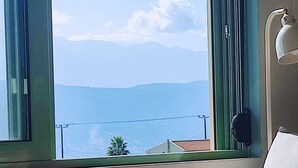 Ein Zimmer mit großem Fenster, das Blick auf das Meer und die Berge gewährt. Es gibt ein Bett mit roter Decke und weißem Kopfkissen, einen Tisch mit einem abstrakten Gemälde in Rot und Weiß sowie eine Lampe auf dem Nachttisch.