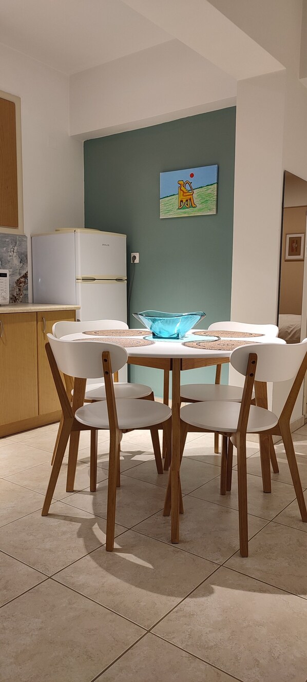 A dining area with a white table and chairs, a refrigerator, and a painting on the wall. - The Garden of Zephyrus – West | Nature & Amazing View (Pithari)