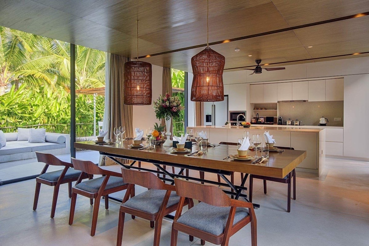 A modern dining area with a long wooden table, six chairs, and a central vase with flowers.