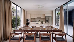 Un'elegante sala da pranzo con un tavolo lungo apparecchiato per sei persone, una cucina sullo sfondo e ampie finestre che offrono vista sull'esterno.