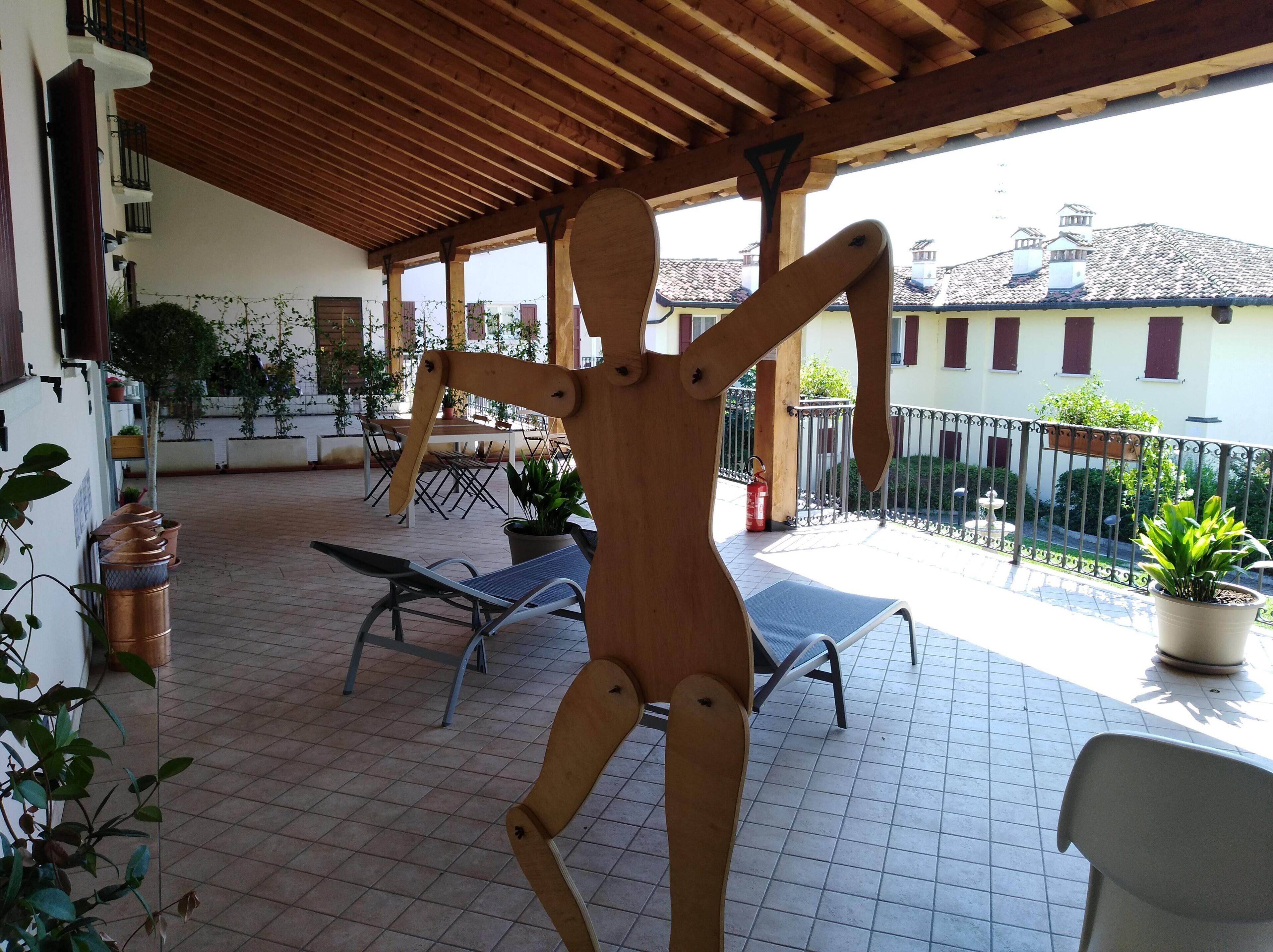 Ein überdachter Patio-Bereich mit Holzbalken, Metallstühlen, Kübelpflanzen und Blick auf Gebäude im Hintergrund.