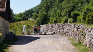Un camino de grava que conduce a un muro de piedra, un pequeño edificio con techo inclinado y un paisaje boscoso al fondo.