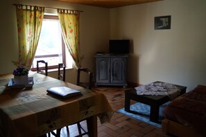 Ein Zimmer mit Esstisch, einer Couch, einem Schrank mit Fernseher und einem Fenster mit Vorhängen.