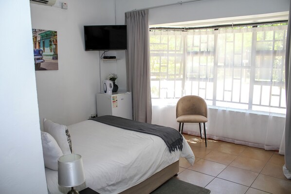 Un dormitorio con cama, televisor montado en la pared, una silla y una ventana con cortinas.