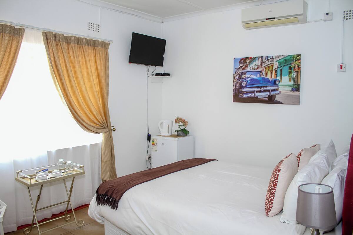 A hotel room with a bed, a TV mounted on the wall, a painting of a car, and a window with curtains.
