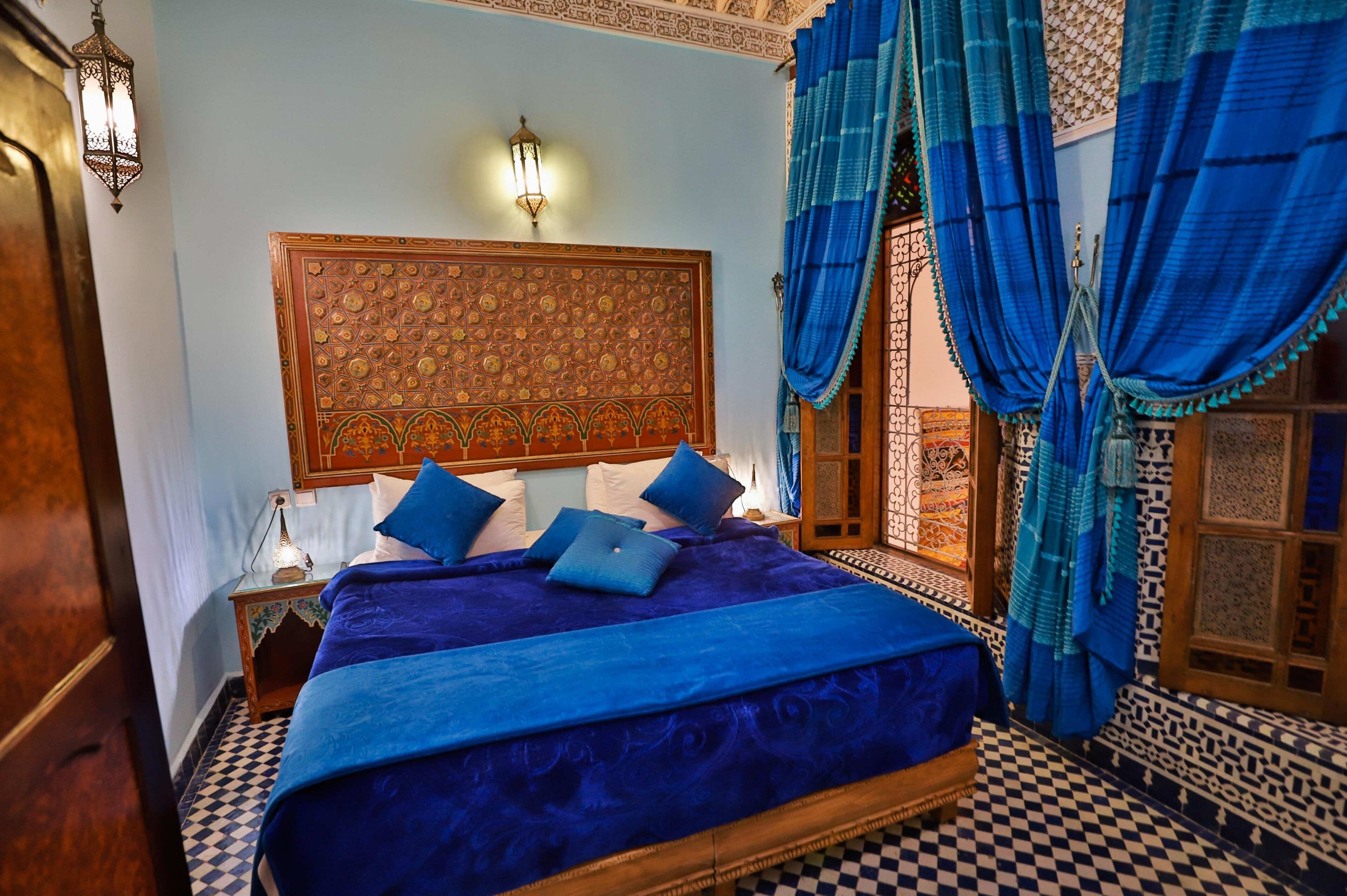 A room with a checkered floor, a bed with blue bedding, a wooden headboard with intricate patterns, and blue curtains.