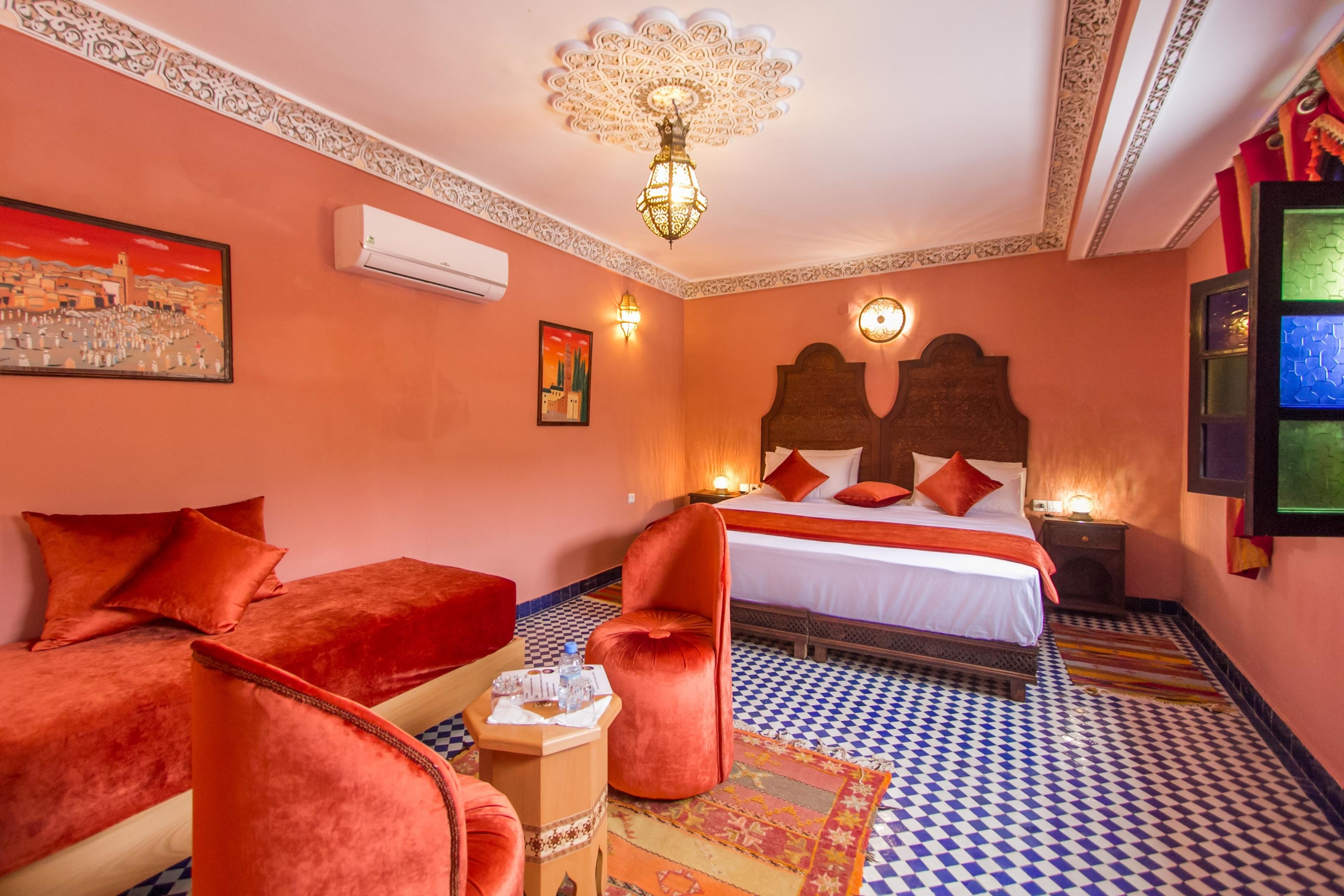 A hotel room with a bed, two red chairs, a small table with a bottle, and a patterned rug.