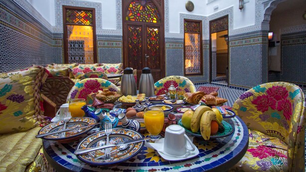 Un comedor marroquí con una mesa redonda dispuesta para el desayuno, que presenta platos, cubiertos y una variedad de alimentos tradicionales marroquíes, incluyendo pan, frutas y un vaso de jugo de naranja.