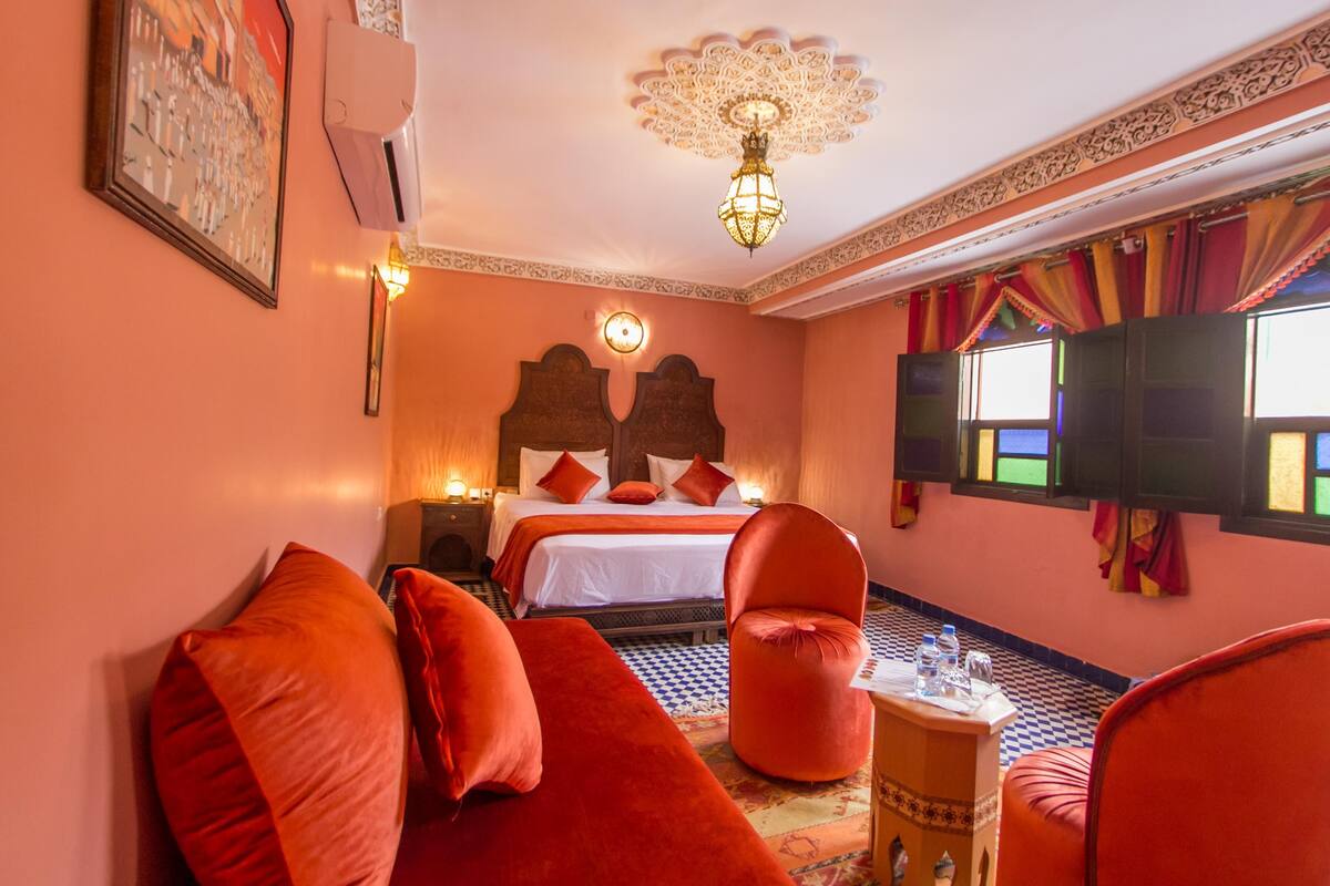 A hotel room with a bed, a sofa, a chair, a small table with water bottles, and a TV.