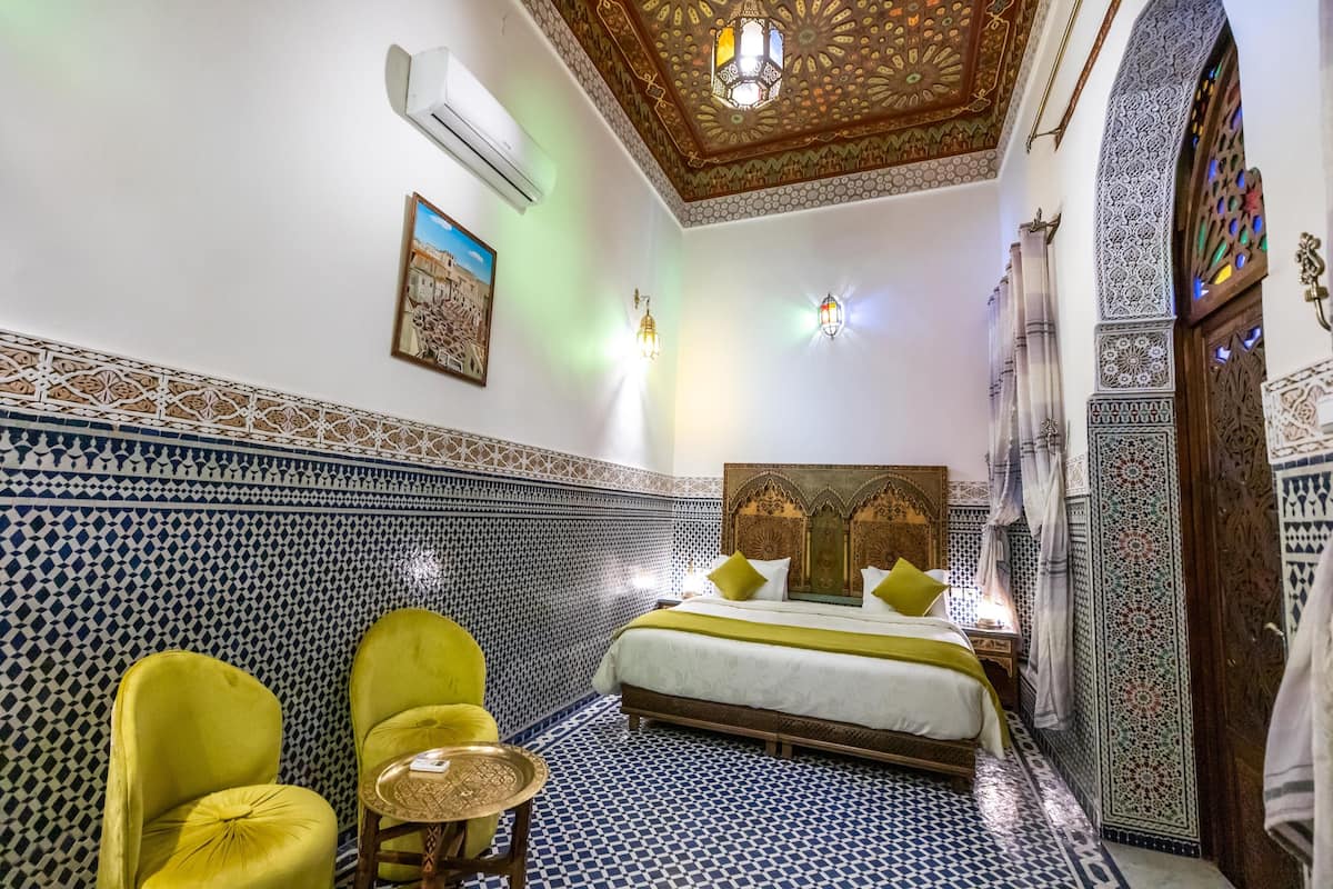 A room with a patterned floor, a bed with a wooden headboard, two yellow chairs, a small round table, and decorative wall tiles.