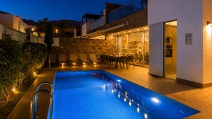 Ein Schwimmbecken mit einer versenkten Sitzecke, ein Essbereich mit Stühlen und Tischen sowie eine durch Glastüren einsehbare Küche.