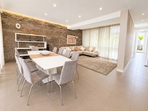 A modern living room with a stone accent wall, a dining area with a white table and grey chairs, a beige sofa, and a large window with curtains. - Spacious 5BR Villa in Costa Adeje with Private Pool (Costa Adeje)