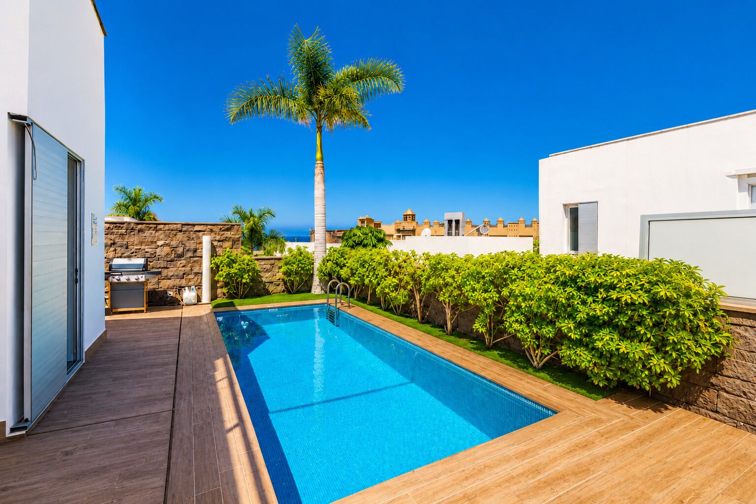 A swimming pool with a wooden deck, a stone wall, and a palm tree.