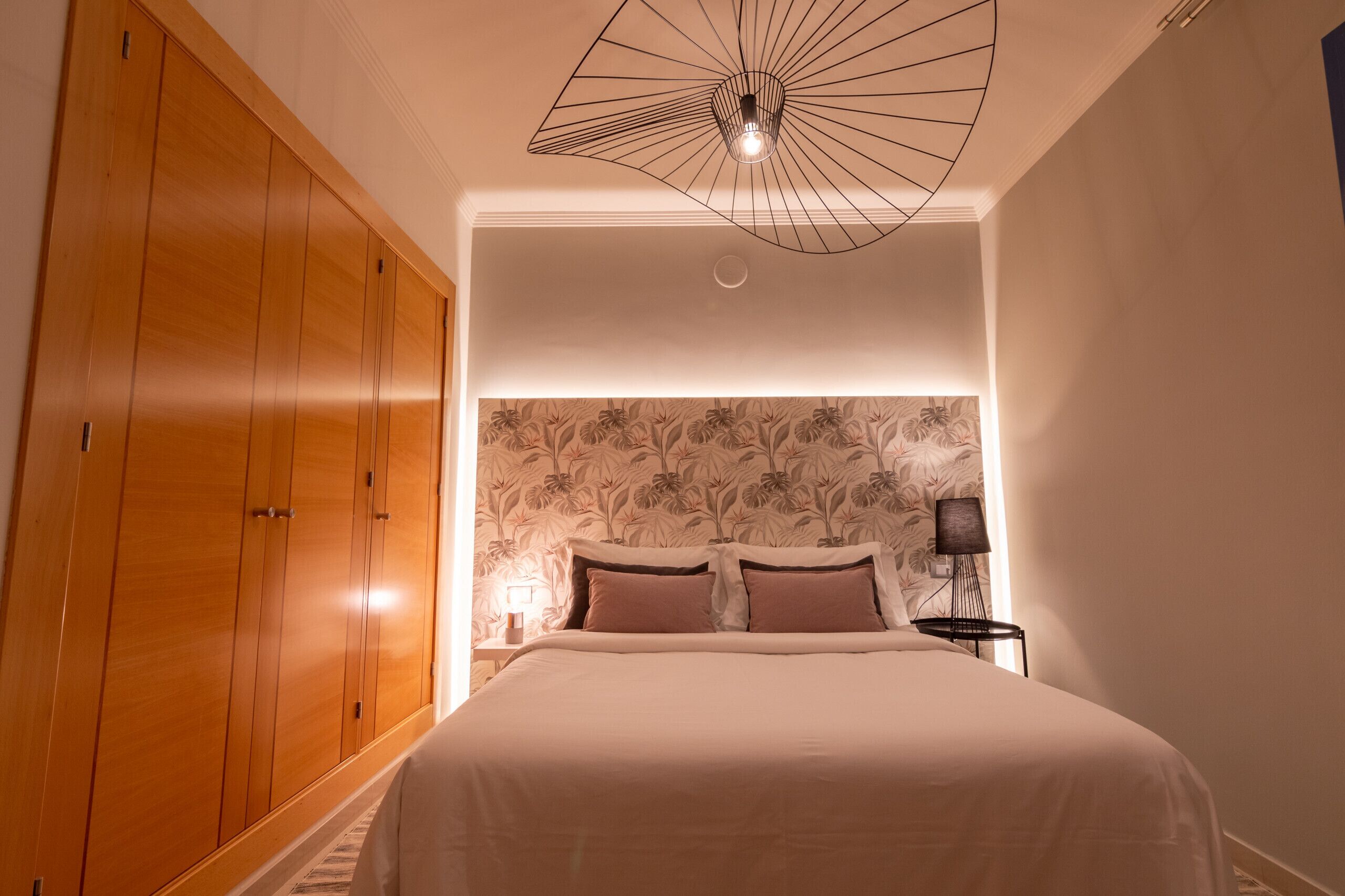 Une chambre avec une grande armoire en bois, un lit doté d’une tête de lit recouverte d’un papier peint à motifs floraux et un luminaire moderne au plafond.